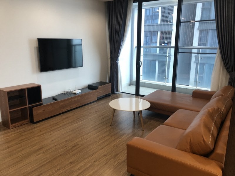 A spacious living area features a comfortable brown leather sofa and a round coffee table. A large flat-screen TV is mounted on the wall. Floor-to-ceiling windows provide ample natural light and access to a balcony.