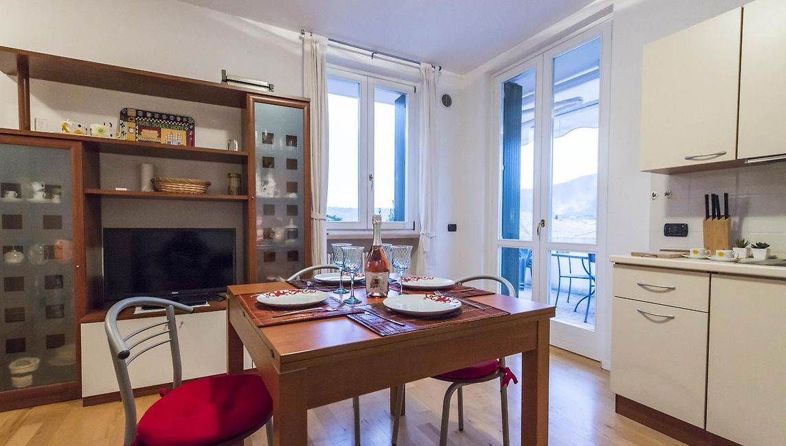 A dining area features a wooden table set for four, accompanied by four chairs. A television sits on a side cabinet, while light pours in through large windows leading to a terrace. A kitchenette is visible, equipped with modern appliances.
