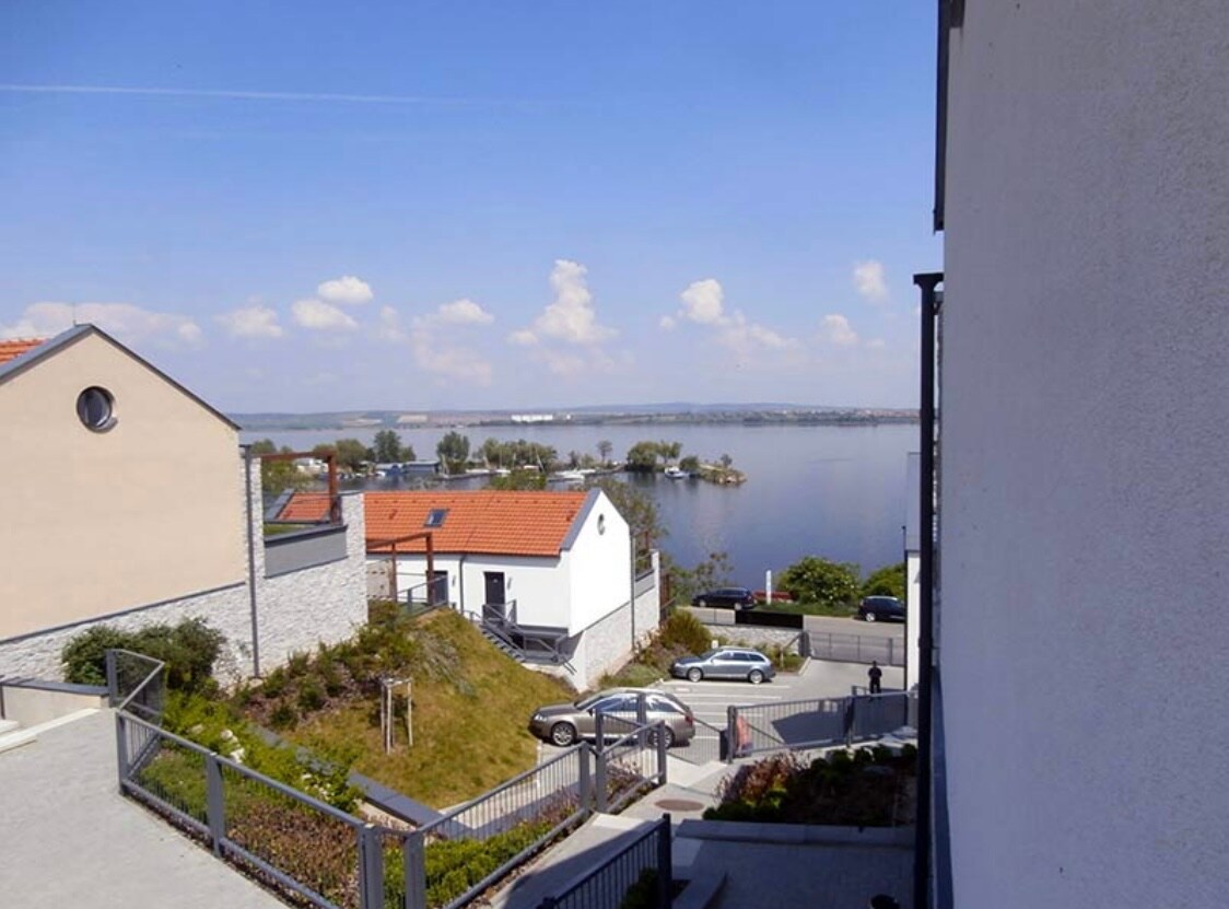 A view of the nearby lake is captured, showcasing calm waters surrounded by greenery. Residential buildings are positioned along the lakeshore, with pathways leading down to the water. The scene is complemented by a clear blue sky and soft, white clouds.