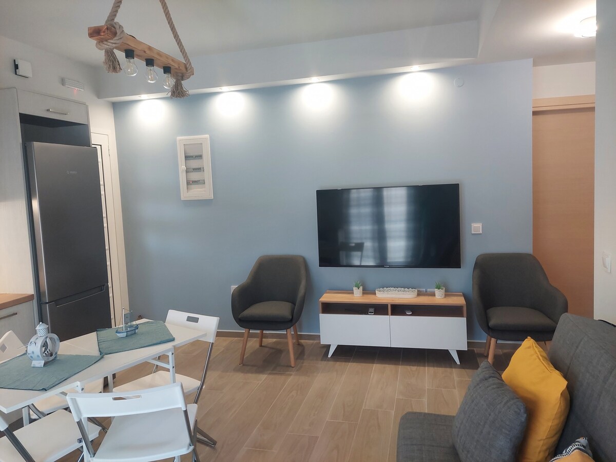 A bright living area features a wall-mounted television, surrounded by two comfortable armchairs and a stylish sofa adorned with a cozy yellow pillow. A dining table with white chairs is positioned nearby, while modern lighting enhances the inviting atmosphere.