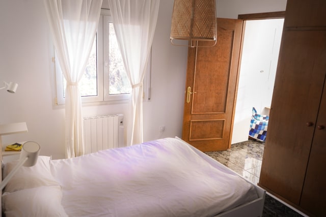 A bright room features a spacious bed dressed in white linens, complemented by soft curtains draping from the windows. Natural light filters through, illuminating the wooden door and the entrance to an adjoining bathroom. The polished floor adds a touch of elegance to the space.