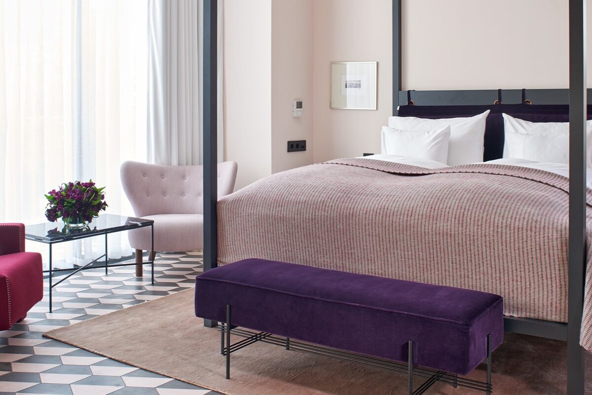 A spacious bedroom is observed, featuring a large four-poster bed with a soft, textured coverlet. A chic purple bench sits at the foot of the bed. Light-colored walls complement a patterned floor. A cozy seating area includes a pink armchair and a glass side table with a flower arrangement.