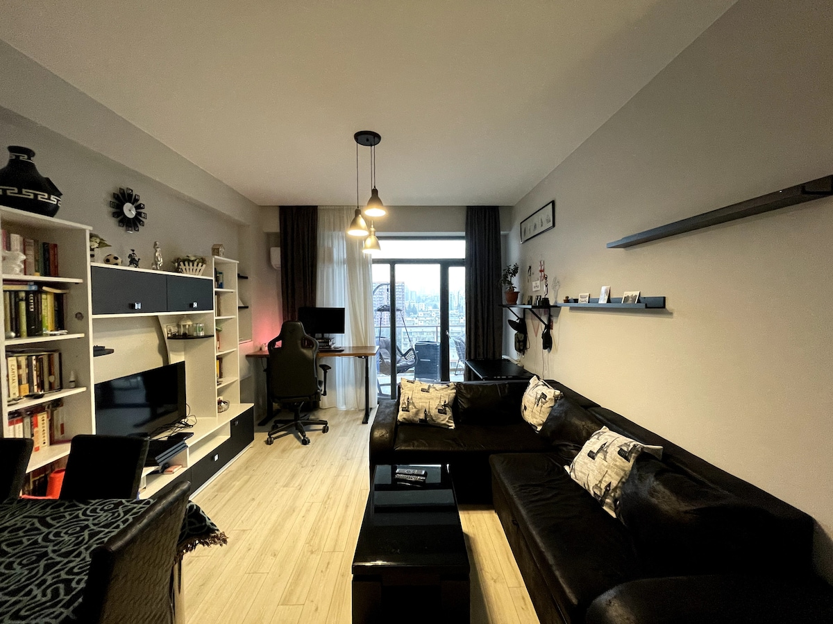 A modern living space features a black sectional sofa and a sleek coffee table. A study area is visible with a desk and chair, while shelves display various decorative items. Large windows allow natural light, offering a view of the city skyline.