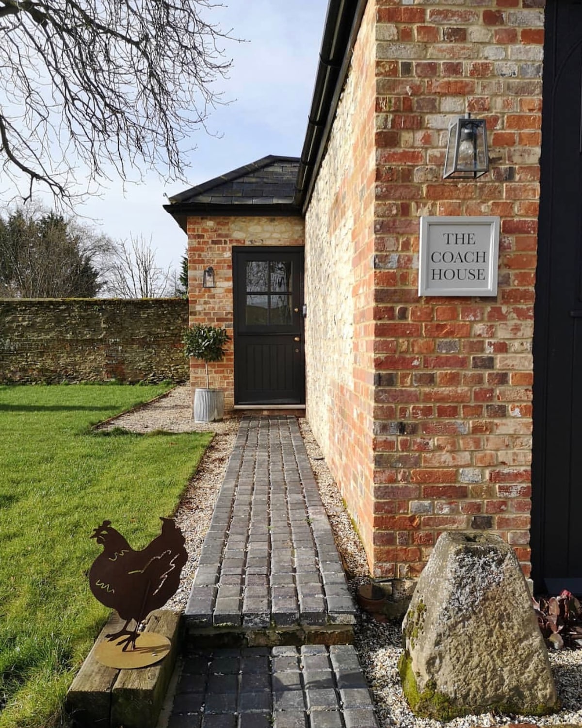 A cobbled path leads to the entrance of The Coach House, framed by a well-kept lawn and low stone walls. A decorative chicken sculpture is positioned near a large rock, and the building's brick façade is complemented by the dark front door.