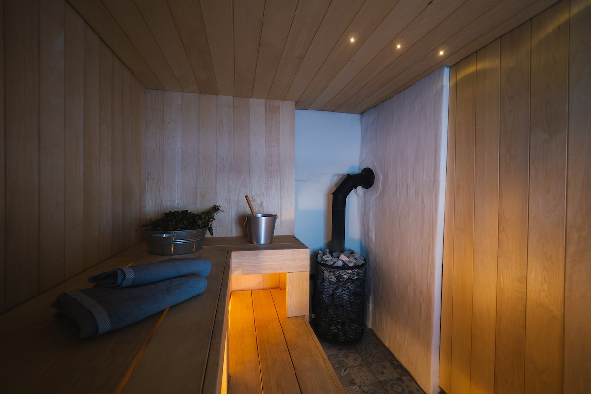 A spacious sauna room features wooden walls and benches, creating a warm and inviting environment. A bucket and ladle are placed beside an aluminum container, while a stove with stones is situated in the corner. Soft lighting enhances the serene atmosphere.