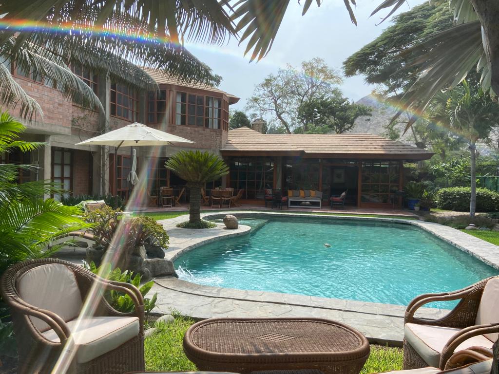 A charming outdoor area features a sparkling swimming pool surrounded by lush greenery. Comfortable seating is arranged nearby, with an umbrella providing shade. The house's large windows reflect the natural light, creating an inviting ambiance for relaxation.