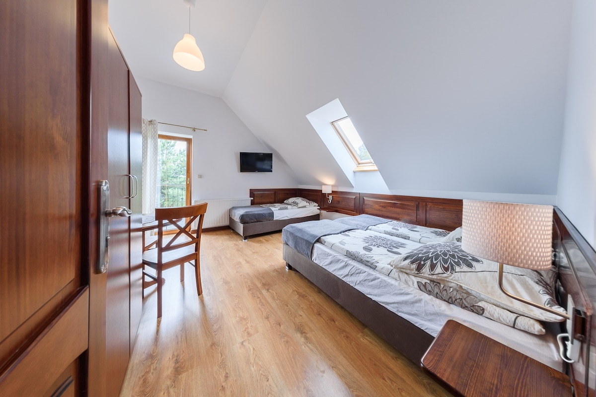 A well-lit bedroom features two spacious beds arranged neatly against wooden paneling. A small desk with a wooden chair is positioned near a window that allows natural light to stream in. A television is mounted on the wall, complementing the cozy atmosphere.