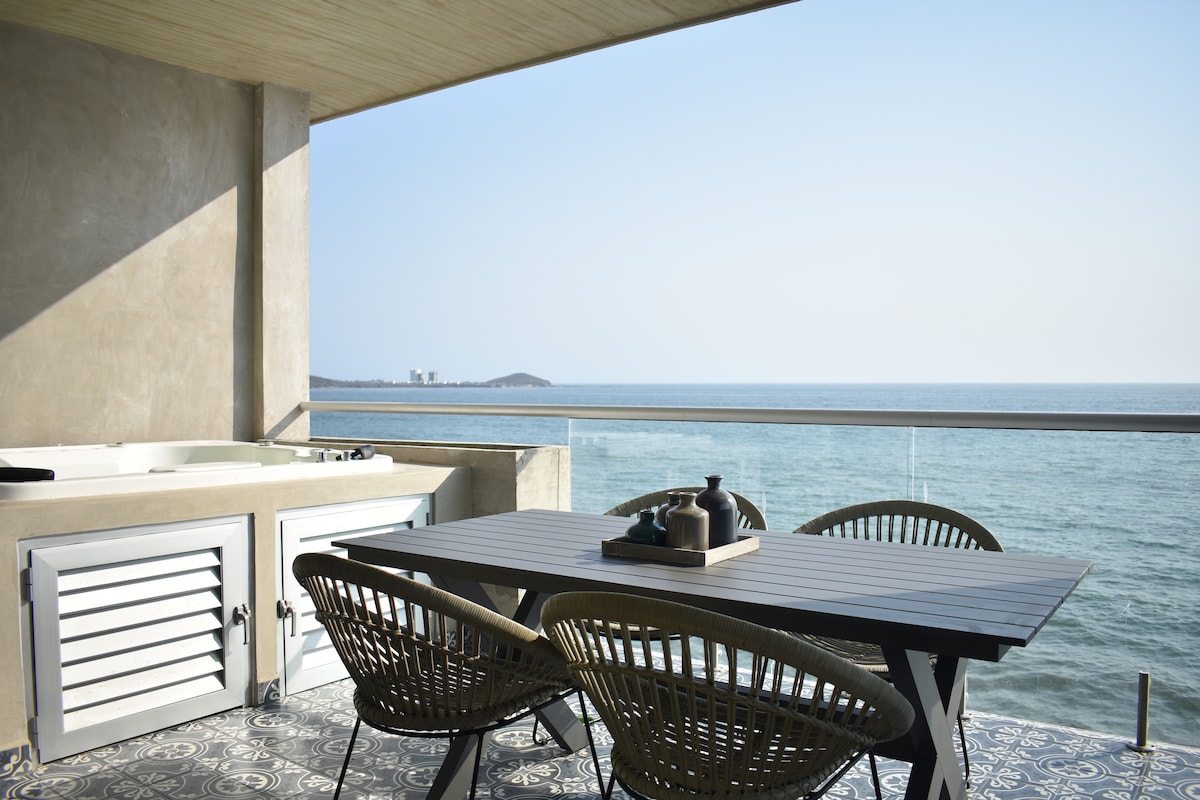 An outdoor balcony features a dining table accompanied by four woven chairs, with a view of the Pacific Ocean. A jacuzzi is positioned adjacent to the table, while the backdrop consists of vast water and a clear sky.