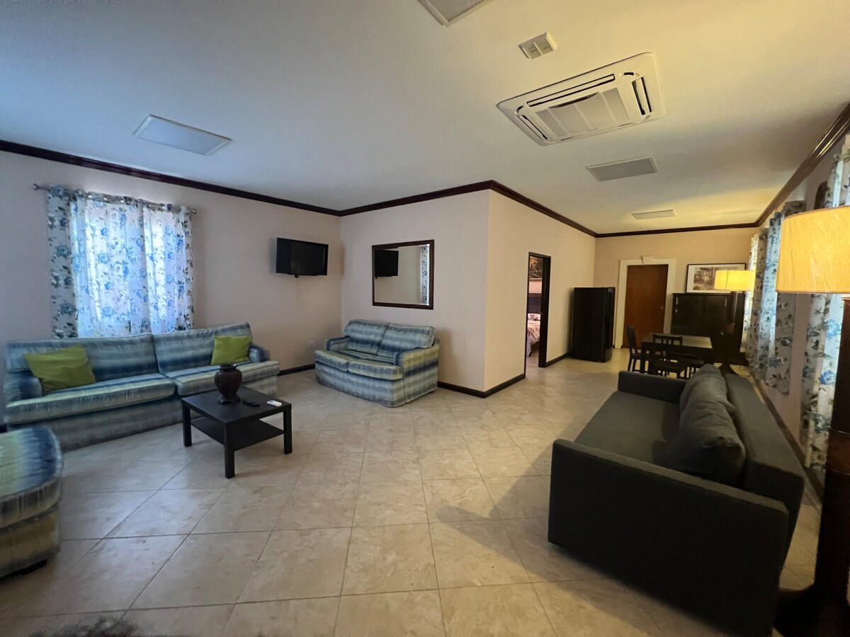 A spacious living area is presented with multiple seating options, including two patterned sofas and a dark sectional. A coffee table is centrally placed, while light filters through floral-patterned curtains. An entertainment unit and dining space are visible, contributing to the room's functional layout.