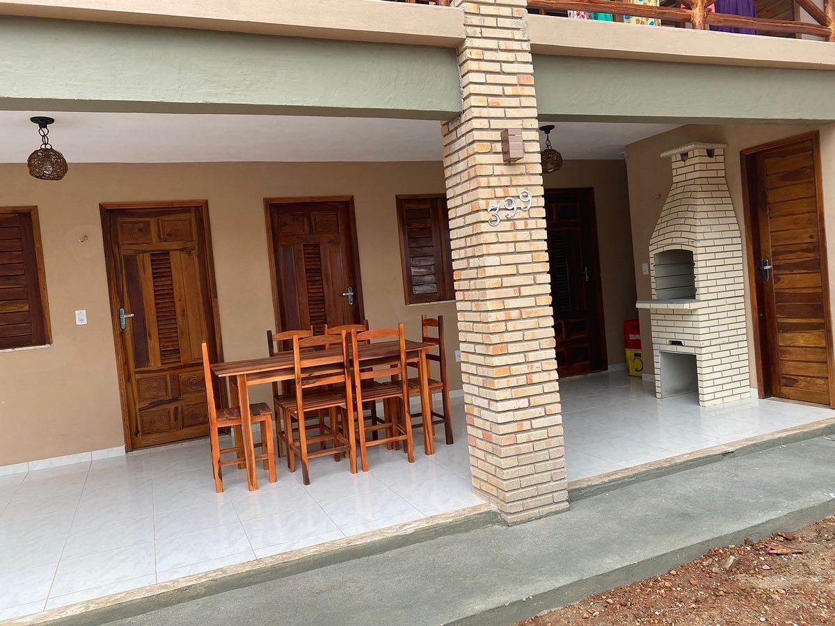 An outdoor dining area is presented with a wooden table and chairs, accommodating several guests. Adjacent to the space, a built-in barbecue grill is visible. The entrance features three wooden doors, leading to private accommodations, framed by textured walls and a smooth tiled floor.