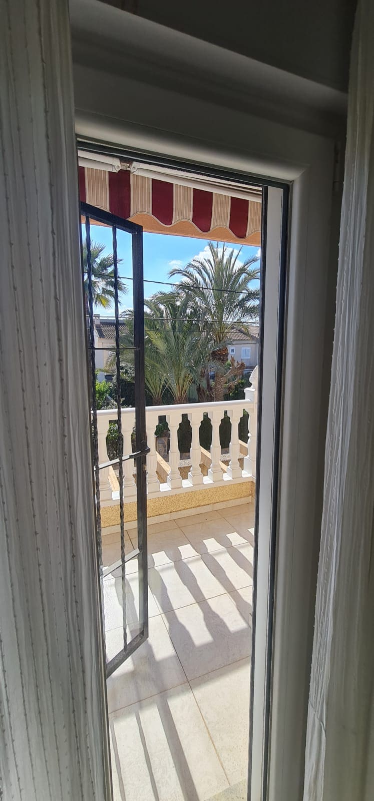 A narrow window frame showcases a view of an outdoor balcony, with a decorative railing and palm trees visible beyond. Stripes of light and shadow fall across the floor, created by the sun filtering through the open door's awning.