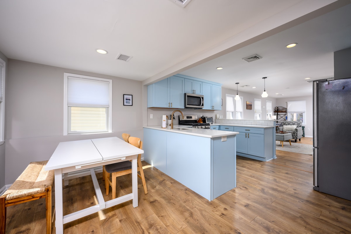 The newly renovated kitchen features light blue cabinetry and stainless steel appliances. A dining table with two chairs sits adjacent to a window, providing natural light. The open layout connects to the living space, showcasing a contemporary design with an inviting atmosphere.
