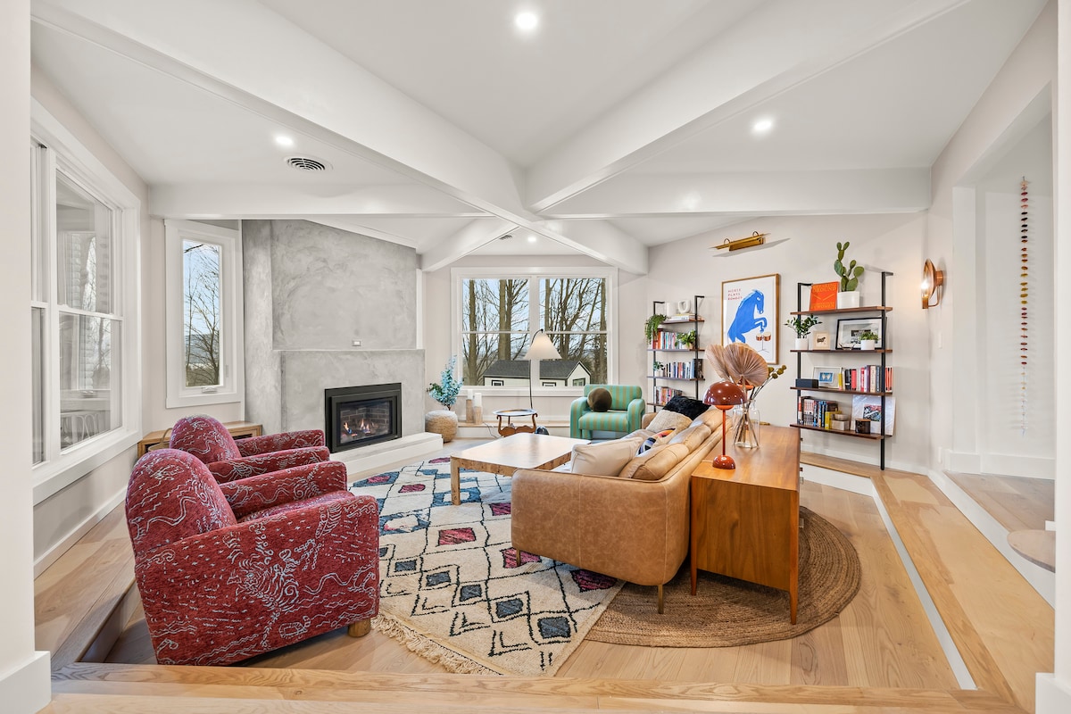 A bright living area features large windows allowing natural light, with views of the outdoors. Cozy seating is arranged around a central coffee table on a patterned rug. Shelving displays books and decorative items, while a fireplace adds a warm touch to the space.