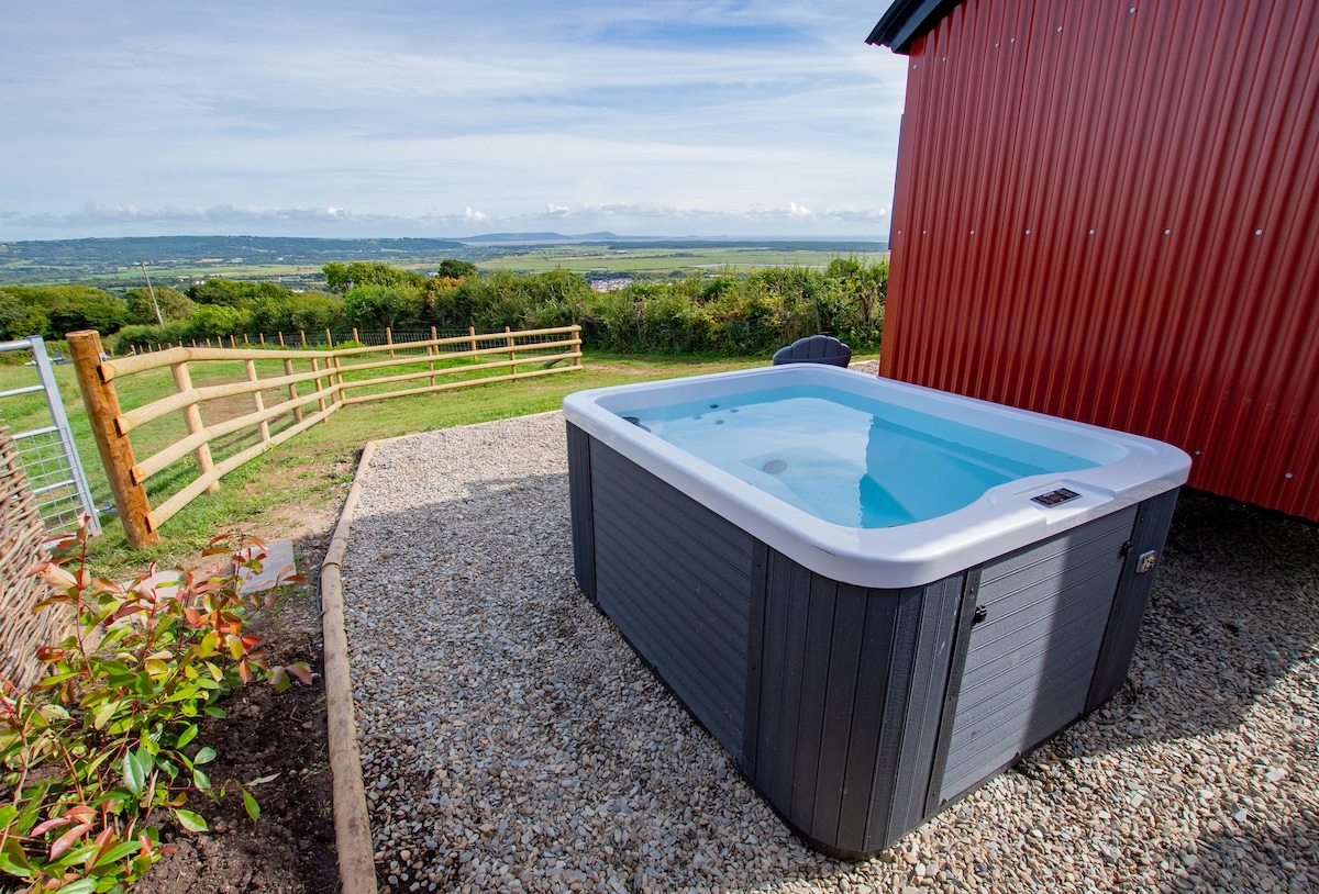 An electric hot tub is positioned on a gravel surface, offering panoramic views of the lush green countryside and distant coastline. The area is surrounded by a wooden fence and includes nearby shrubs, creating a serene outdoor space.
