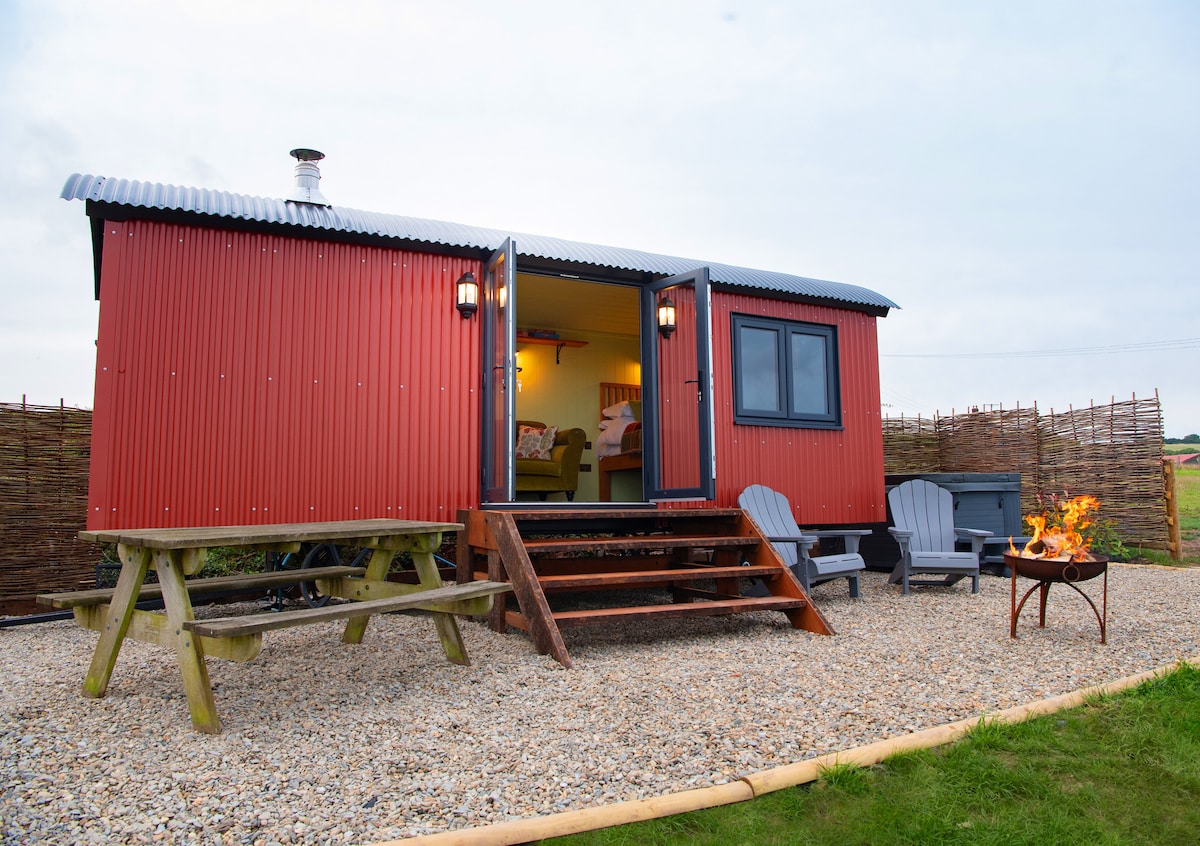 The exterior of a traditional Shepherd's Hut is showcased, featuring a vibrant red corrugated facade. Steps lead up to double doors, revealing a cozy interior. A fire pit with seating and a picnic table are positioned within a gravel area, contributing to the outdoor relaxation space.