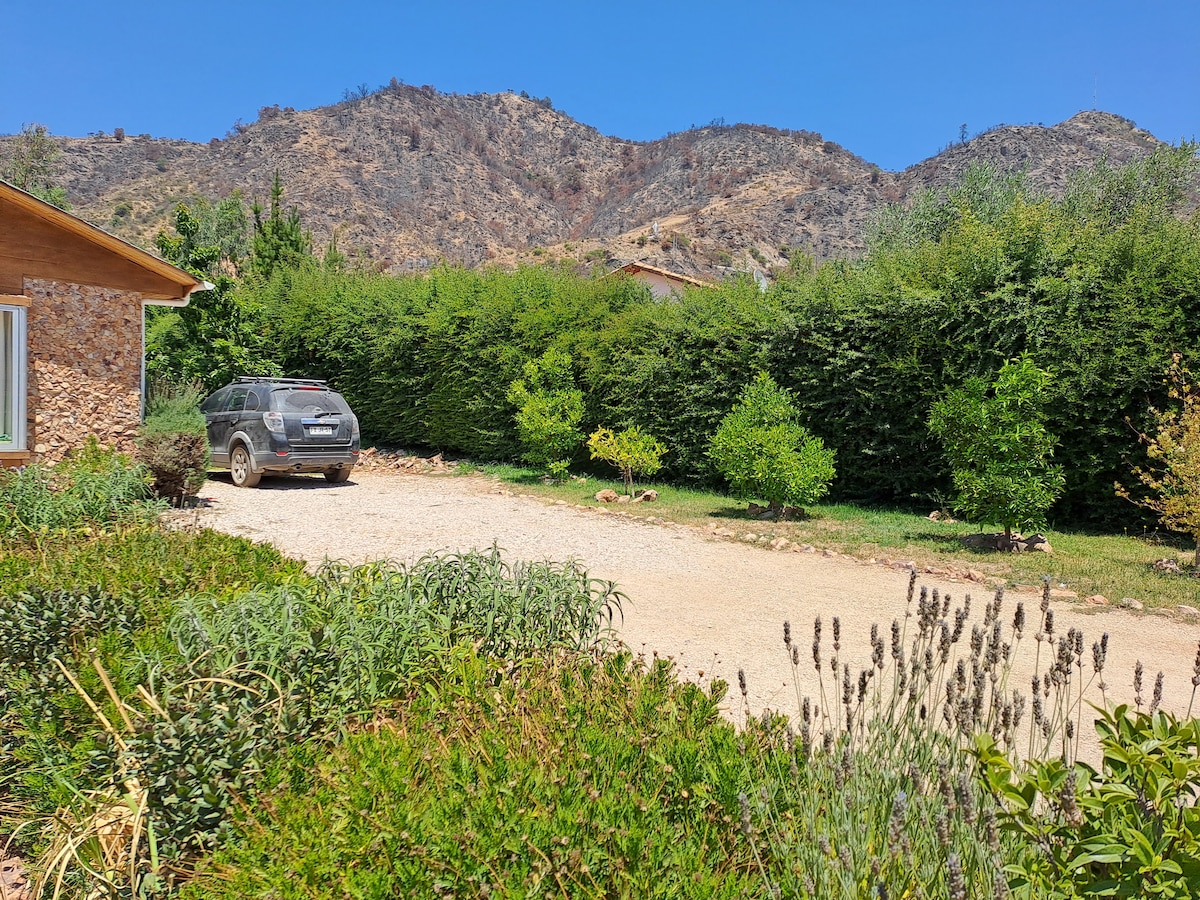 A gravel driveway leads to a house, bordered by lush green hedges and trees. The background features dry, mountainous terrain under a clear blue sky. Various plants, including lavender, are visible in the foreground, contributing to the rural landscape.