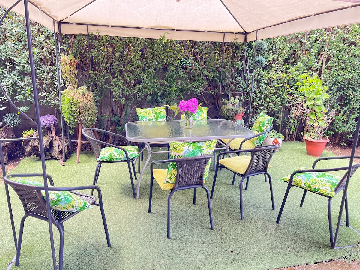 An outdoor seating area is covered by a canopy, featuring a glass table surrounded by six chairs with vibrant tropical-patterned cushions. Potted plants and lush greenery create a serene backdrop, adding to the inviting space for relaxation and gatherings.