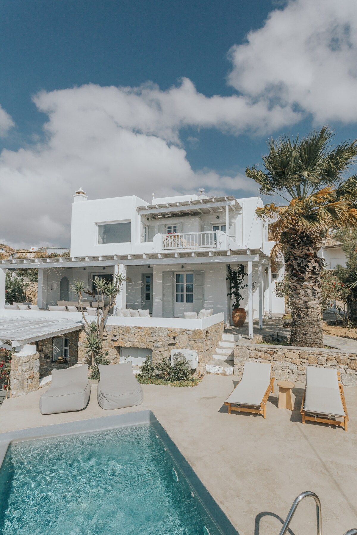 The villa showcases a modern white exterior with multiple balconies and large windows, situated against a backdrop of palm trees. A private swimming pool reflects the sky above, while lounge chairs are arranged nearby on a stone patio.