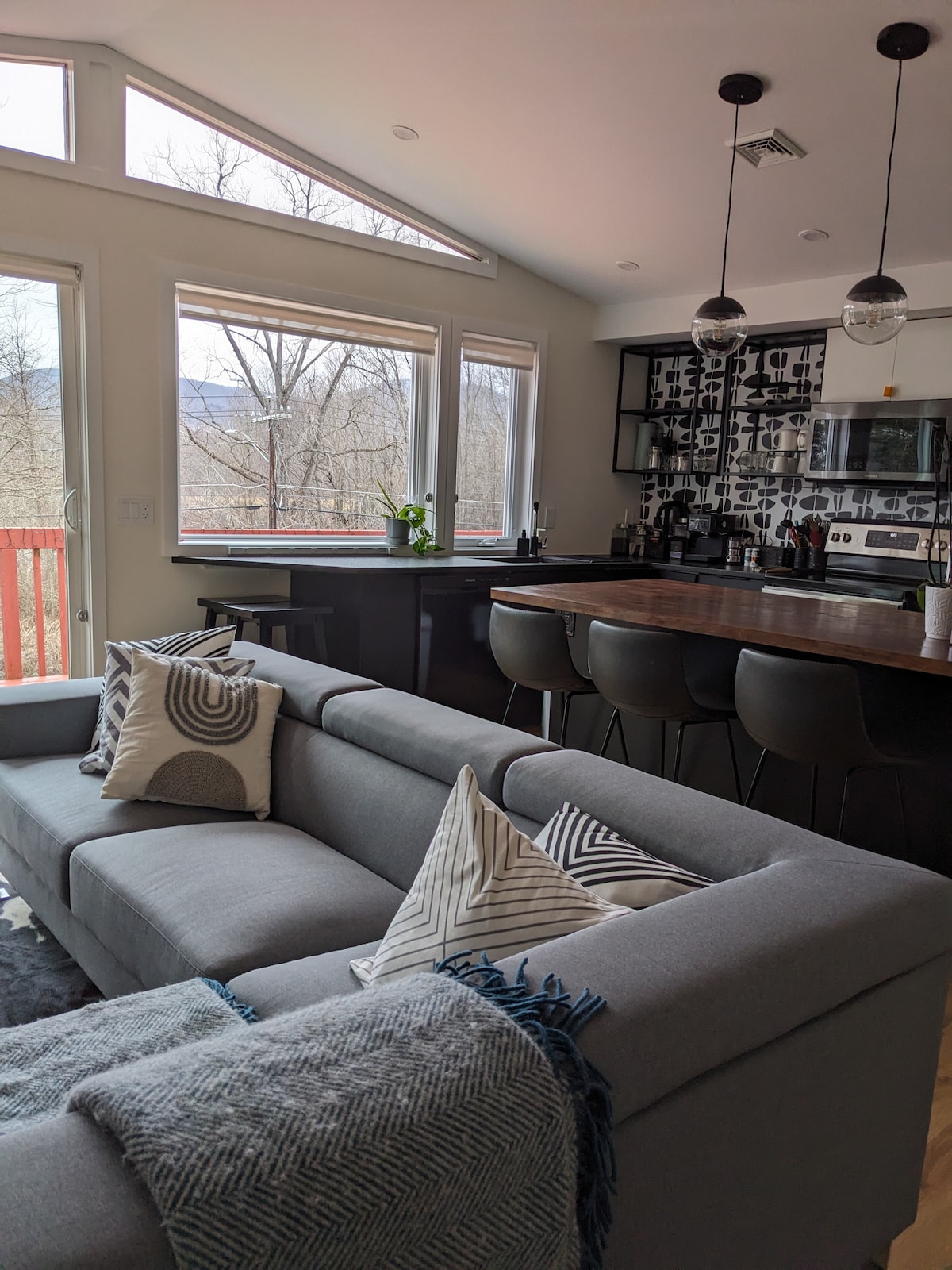 A modern living area is showcased with a grey sofa adorned with patterned pillows. A large wooden counter with high stools is visible, alongside a kitchen featuring sleek cabinetry. Expansive windows allow natural light to fill the space, framing views of the outdoors.
