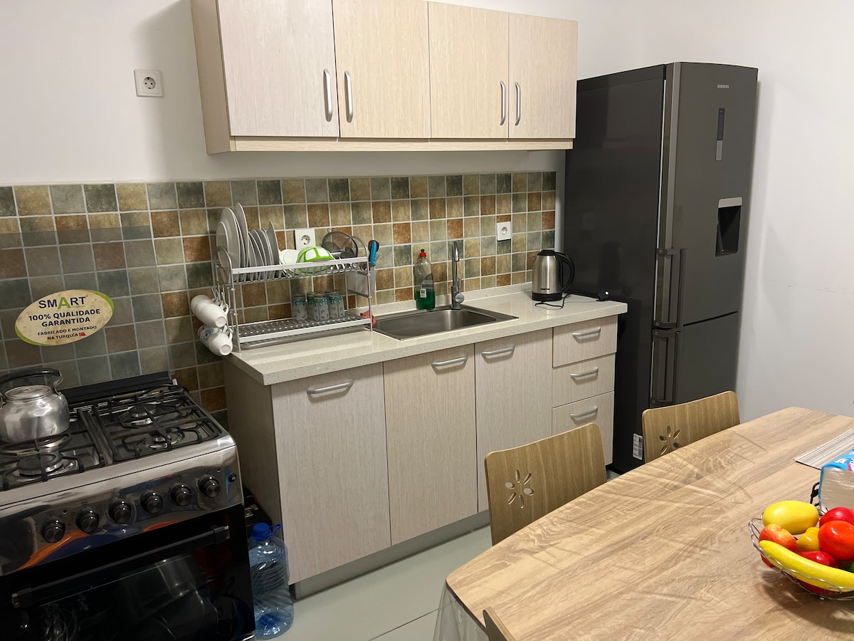 A well-equipped kitchen features a stove, sink, and refrigerator. Wooden cabinets and a tiled backsplash add functionality and style. A dining table with chairs is positioned nearby, and a bowl of fresh fruit is displayed at its center.