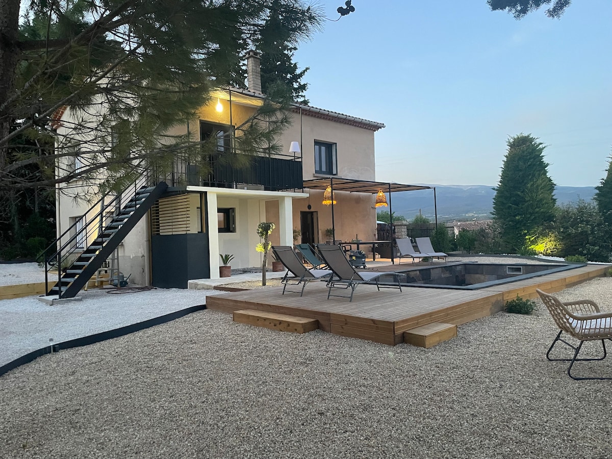 The renovated house is set within a landscaped garden, featuring a spacious wooden terrace surrounding the swimming pool. Outdoor seating is provided on the terrace, with steps leading up to a balcony accessible from the upper level of the home.