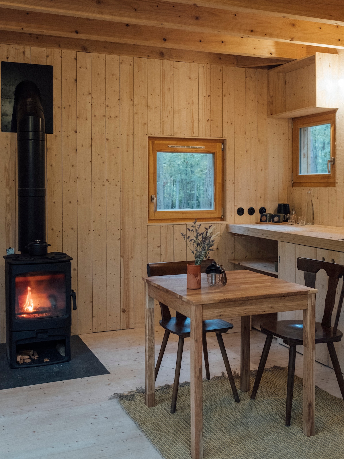 A cozy dining area features a wooden table with two matching chairs, complemented by a small vase on the table. A black wood stove adds warmth to the space, with large windows allowing natural light to illuminate the pine-paneled walls.