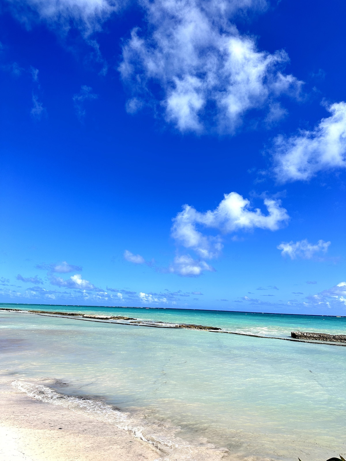 A serene beach scene features soft white sand that meets clear turquoise waters. The horizon showcases gentle waves under a bright sky adorned with scattered white clouds. The tranquil environment invites relaxation and appreciation of nature's beauty.