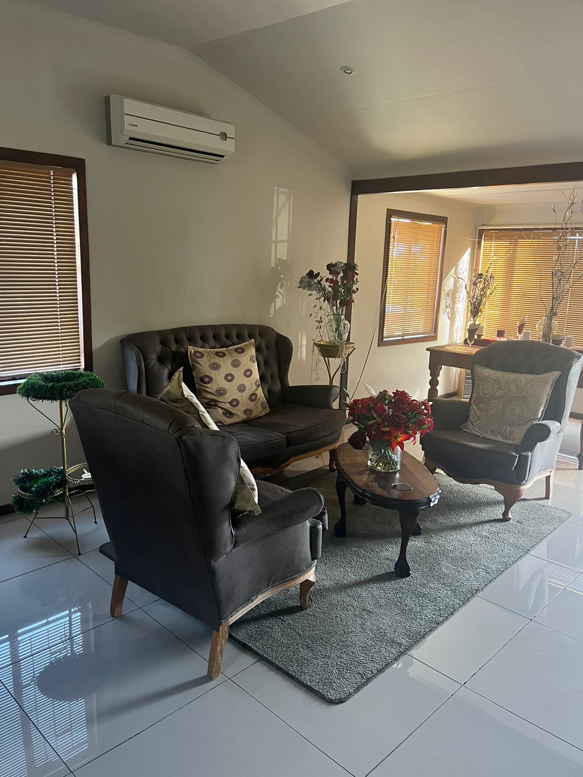 A cozy living area is furnished with a dark leather sofa and an elegant armchair adorned with decorative cushions. A round wooden table holds a vibrant floral arrangement, while natural light streams through the large windows, highlighting the warm tones of the decor.