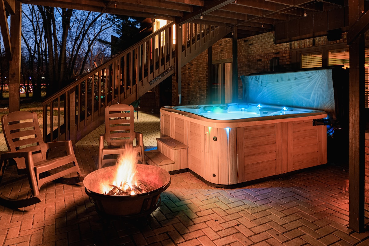 A wooden hot tub is positioned under a deck, surrounded by a brick patio. Soft lighting illuminates the area. Nearby, a fire pit with glowing embers is accompanied by four wooden chairs, creating a relaxing outdoor space for gathering.