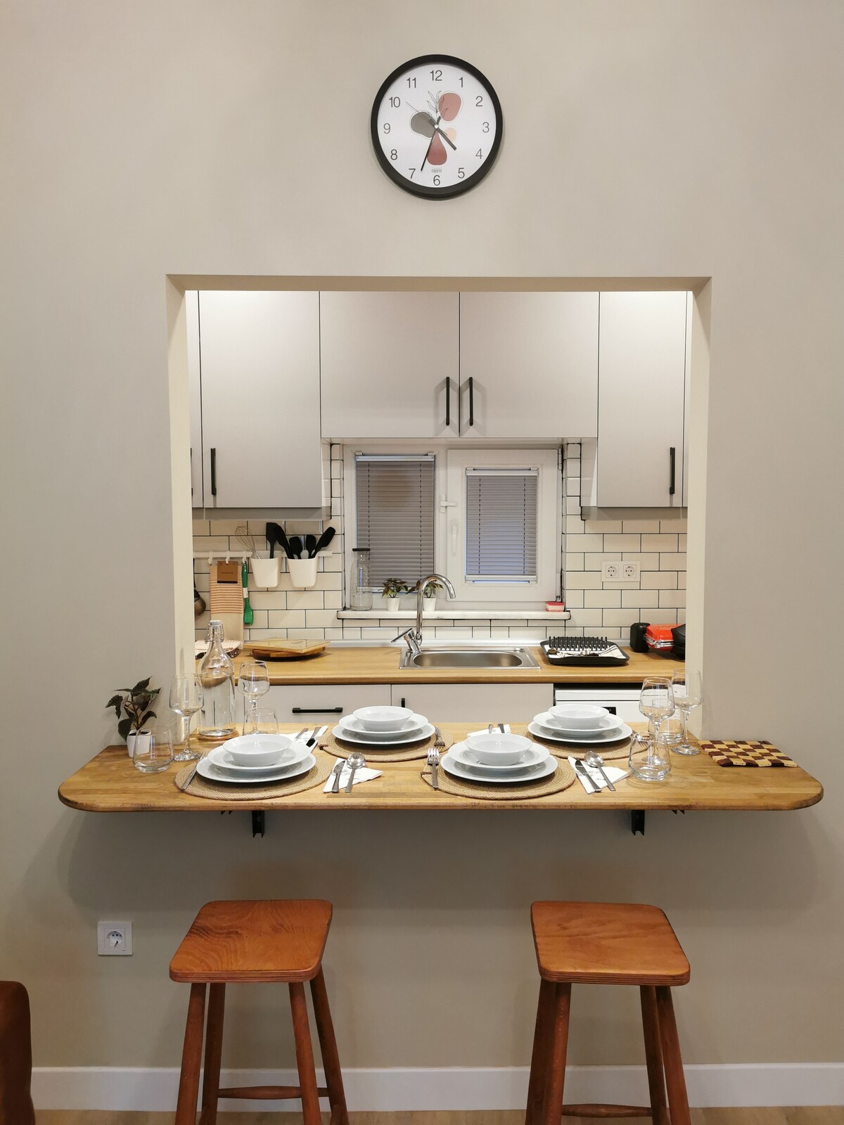 A functional kitchen area is designed with a shallow bar counter, set with plates, cutlery, and glassware. An open window provides views into the kitchen, which is equipped with cabinetry and modern appliances. A wall clock is mounted above the counter.