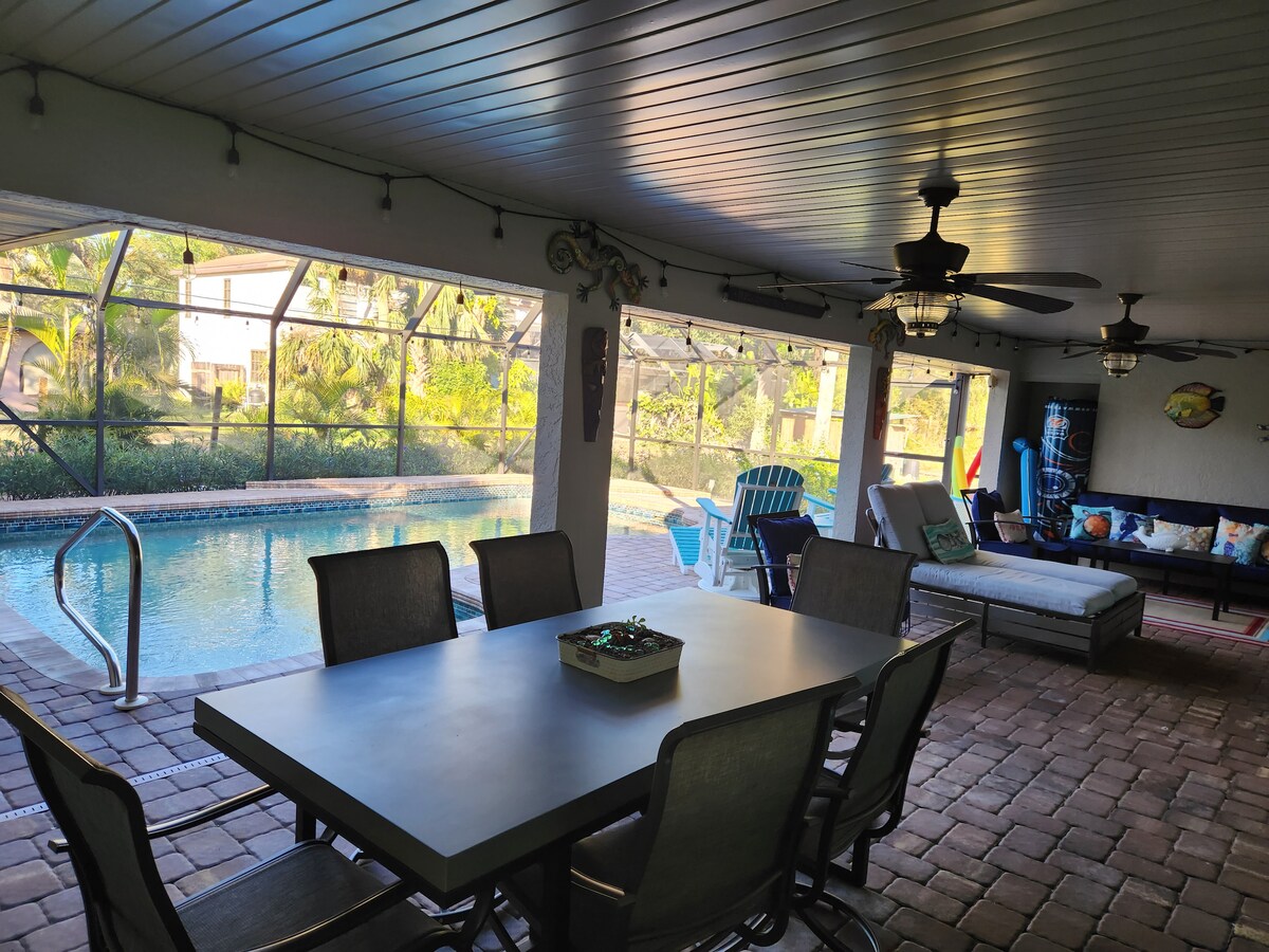 A covered patio area is shown, featuring a large dining table surrounded by chairs, and a lounge area with comfortable seating. A saltwater pool is visible beyond, framed by greenery and a screened enclosure, providing a blend of indoor and outdoor relaxation spaces.