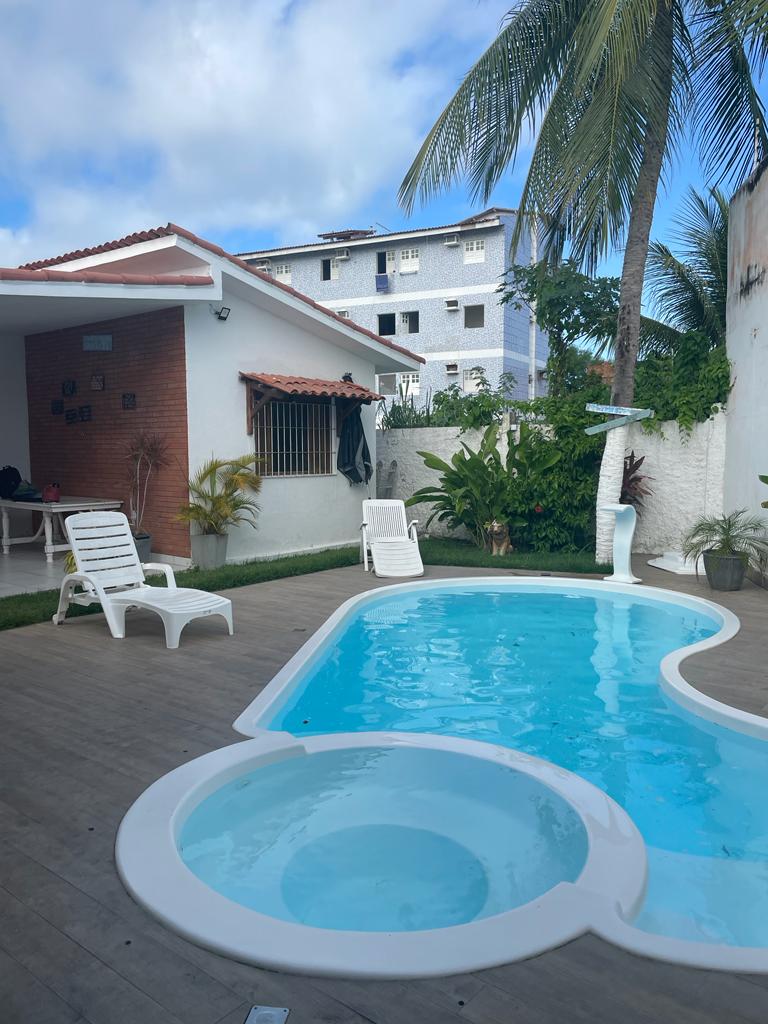 A swimming pool is surrounded by a wooden deck, featuring a separate hot tub area. Lounge chairs are positioned nearby, accompanied by tropical plants and palm trees. A single-story building is visible in the background, offering a relaxed space for outdoor enjoyment.