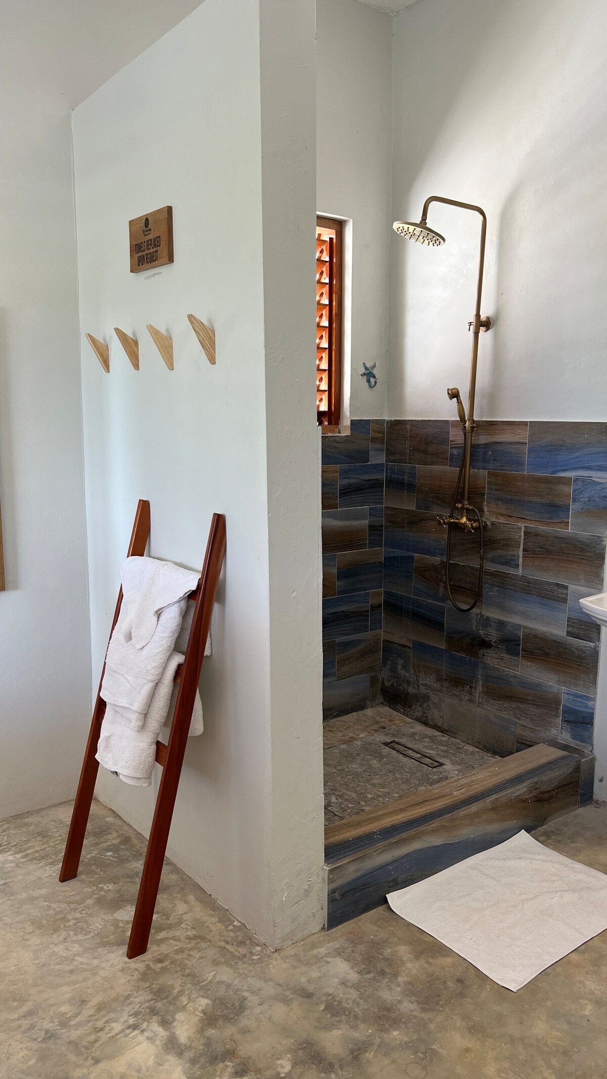 A stylish shower area features a gold showerhead mounted on a tiled wall, combining earthy hues. A wooden ladder displays neatly folded towels. A small mat lies on the floor, complementing the neutral tones of the cement space.