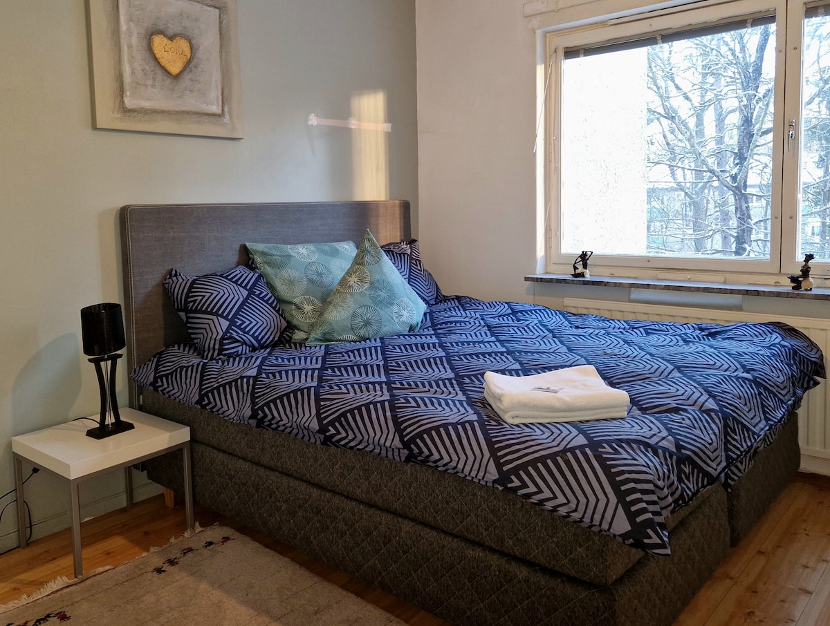 A spacious bedroom features a large bed with patterned bedding and several pillows. A small bedside table holds a lamp. Natural light enters through a window that provides a view of trees outside, enhancing the peaceful ambiance of the room.