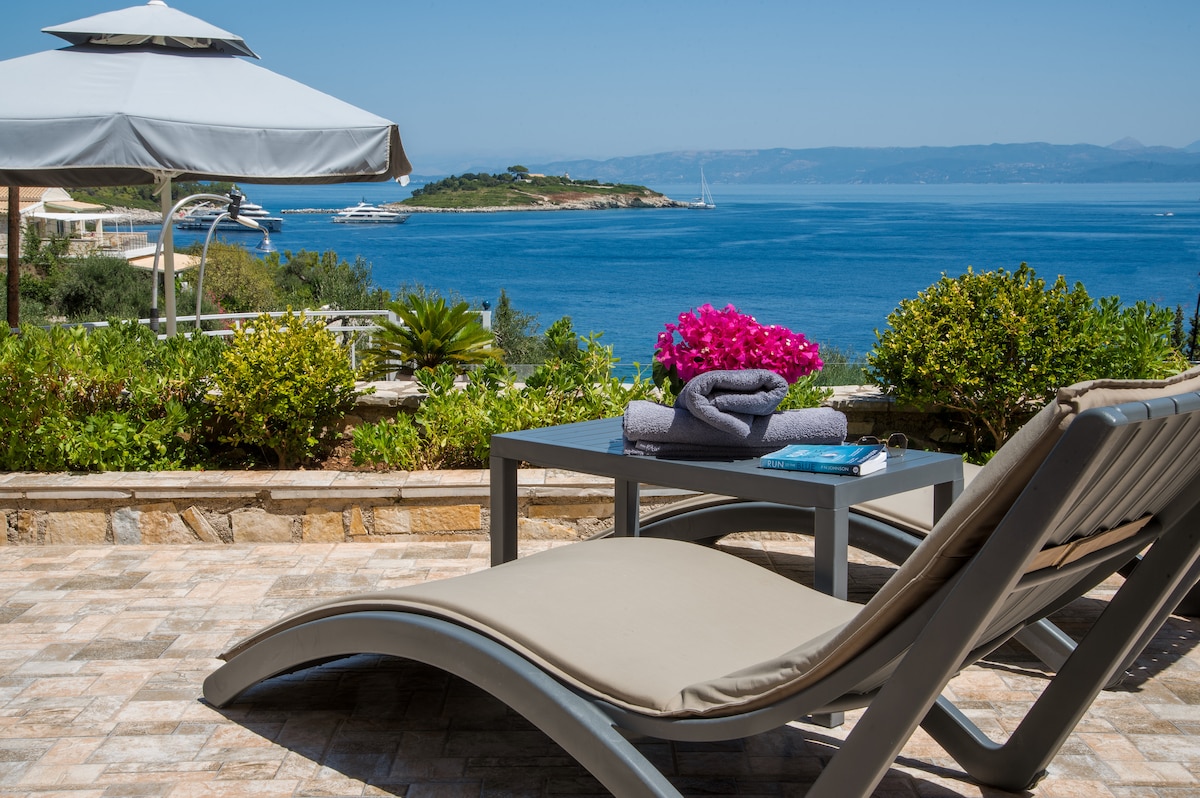 A comfortable sun lounger is positioned on the terrace, overlooking tranquil waters of the Ionian Sea. Bright pink flowers add a pop of color to the scene. A small table holds a stacked towel and a book, with lush greenery framing the picturesque view.