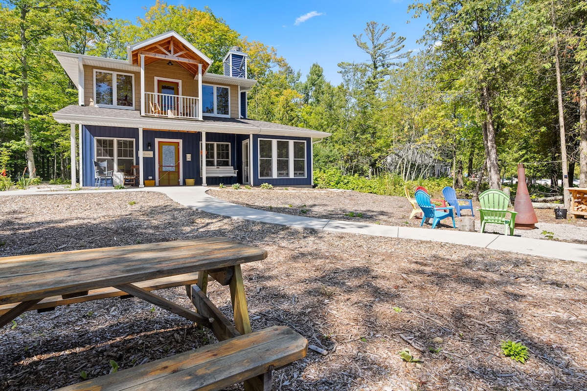 A welcoming two-story cottage is surrounded by trees and features a spacious front yard. A wide concrete pathway leads to an entrance. Colorful outdoor seating is arranged near a fire area, inviting relaxation and social gatherings in the natural setting.