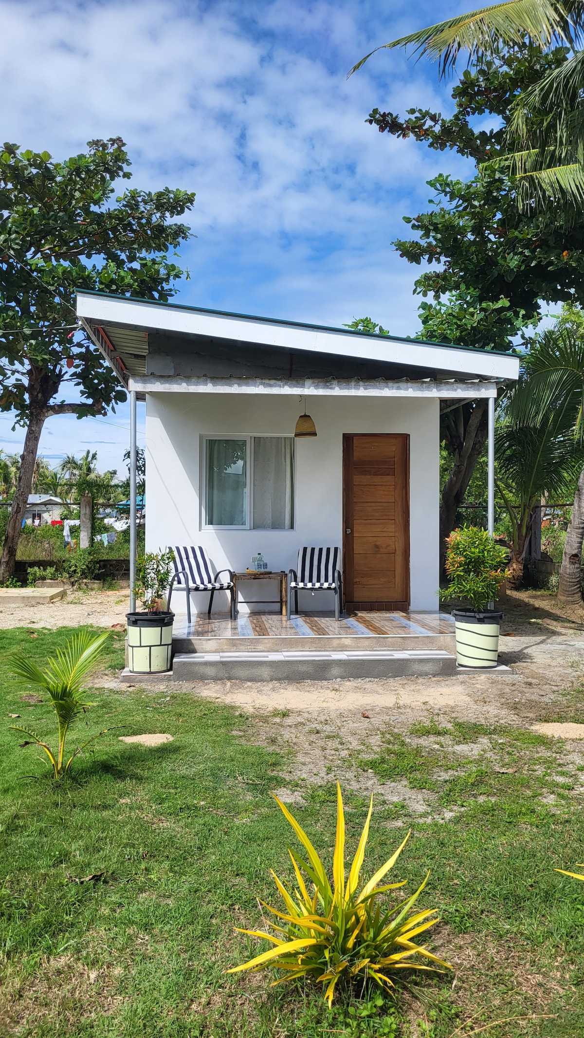 A small, modern bungalow features a welcoming entrance with a wooden door and a covered porch. Two striped chairs are positioned on the porch, complemented by potted plants. Lush green grass and palm trees surround the property, enhancing the outdoor setting.