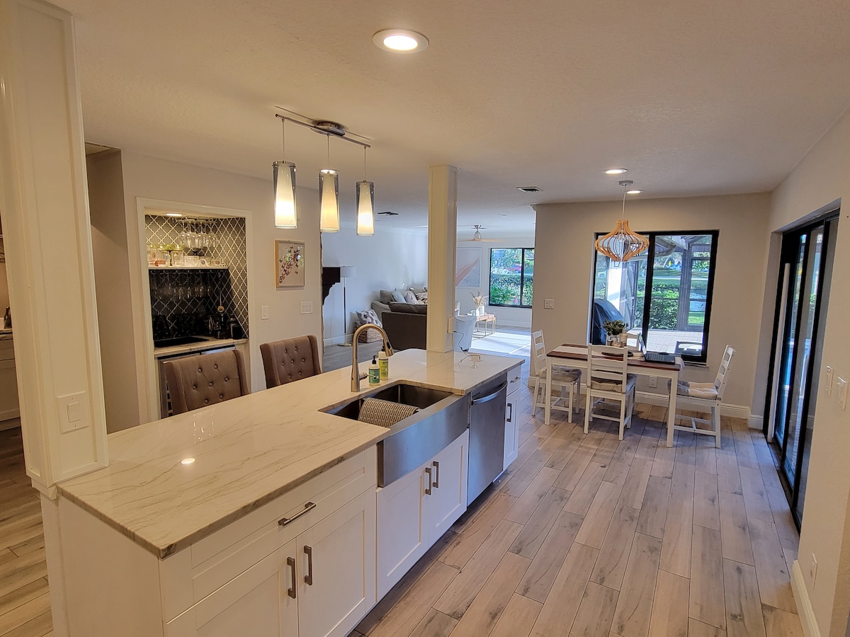 A spacious kitchen features a central island with a sink. Modern pendant lights hang above, illuminating the area. To the side, a dining space with a table and chairs is visible. Large windows invite natural light and showcase views of the outdoor area.