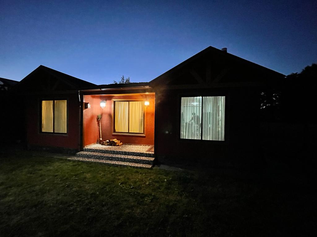 The exterior of the cabin is illuminated at night, showcasing a warm entryway with large windows. A soft glow from the porch lamp highlights the stone steps, while the darkening sky enhances the welcoming appearance of the home.