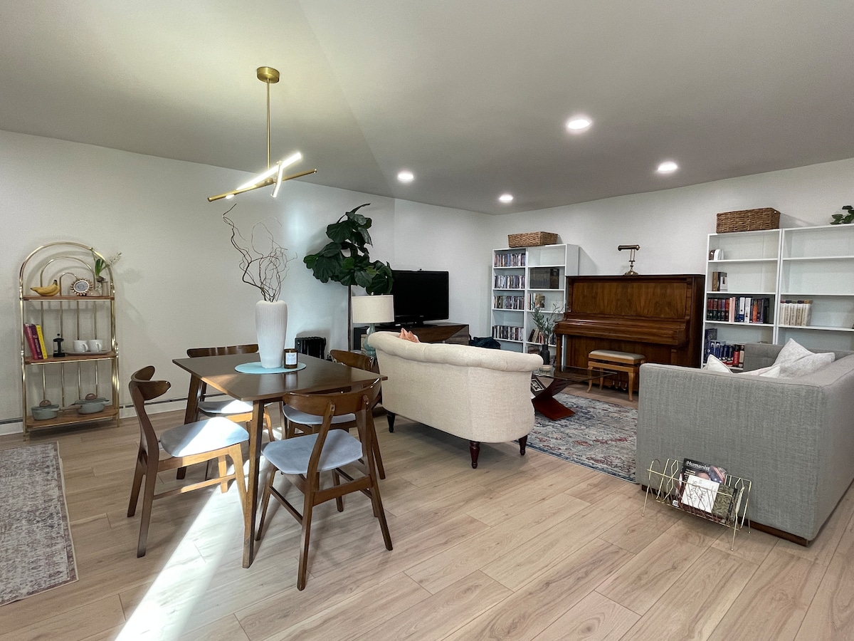 An open-concept living space showcases a dining area with a round table and four chairs. A comfortable couch faces a media unit and a piano. Bookshelves line the walls, while natural light highlights indoor plants, creating a bright and inviting atmosphere.