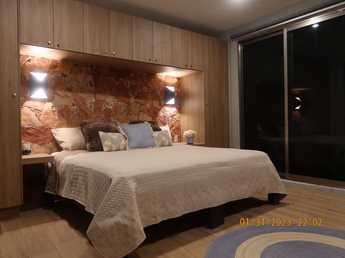 A spacious sleeping area features a neutral quilt covering a large bed. Soft lighting is provided by built-in wall sconces flanking a textured stone wall. A round rug lies on the wooden floor, and a large window allows for natural light.
