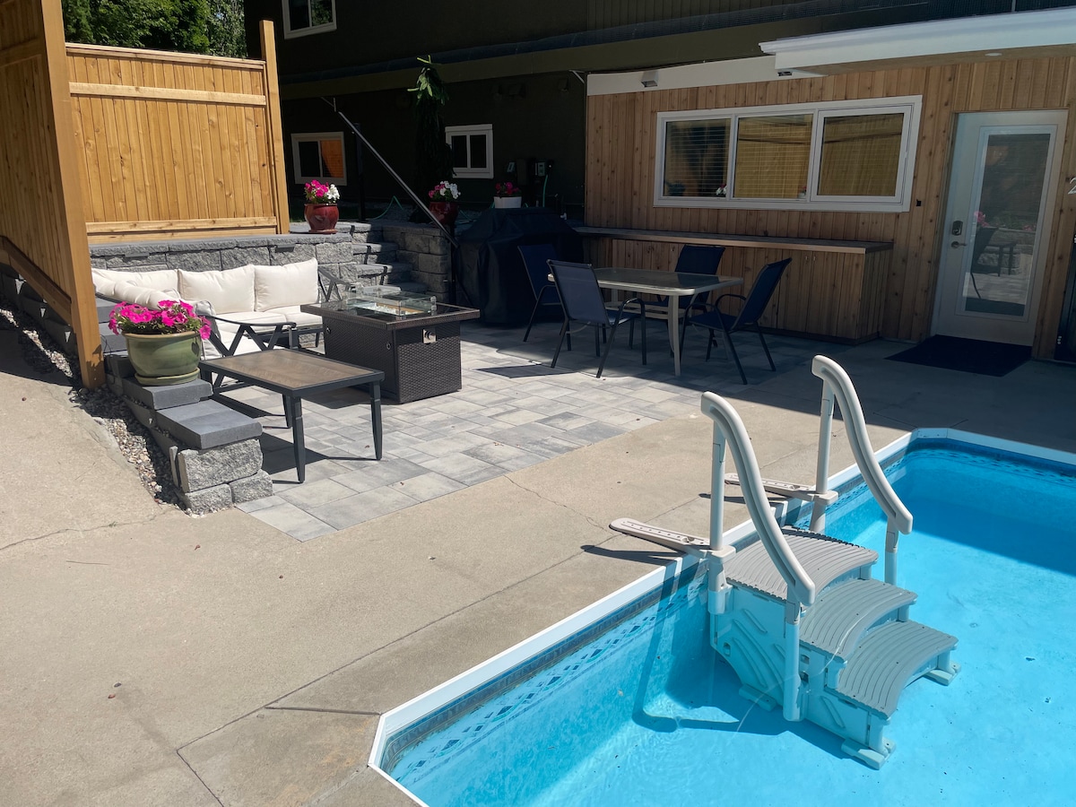 A pool area features a heated salt water pool with steps for easy access. Surrounding the pool is a paved patio that includes outdoor furniture, a dining table, and a barbecue grill. Colorful flower pots add a touch of greenery to the space.