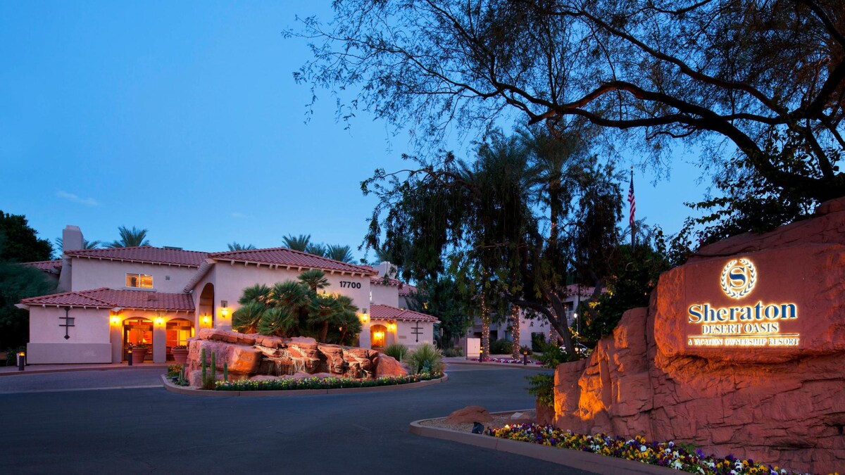 The entrance of Sheraton Desert Oasis is highlighted by a warm-toned stone sign against a backdrop of lush landscaping and palm trees. The resort building features a rustic design, with terracotta roof tiles and desert foliage, creating a welcoming atmosphere at dusk.