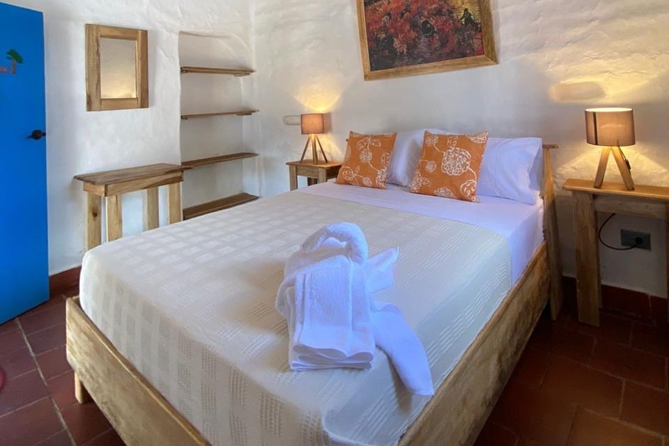 A cozy bedroom features a spacious bed dressed in light bedding, complemented by decorative pillows on the headboard. Wooden bedside tables with lamps flank the bed, while a mirror and open shelving provide additional functionality against the whitewashed wall.
