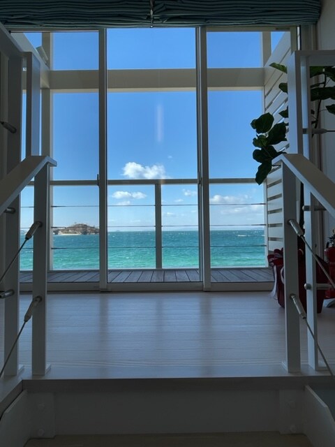 A spacious entrance is highlighted by large glass doors that provide an unobstructed view of the ocean. Natural light floods the area, illuminating the sleek wooden flooring. Green plants are positioned nearby, adding a touch of nature to the coastal scene.