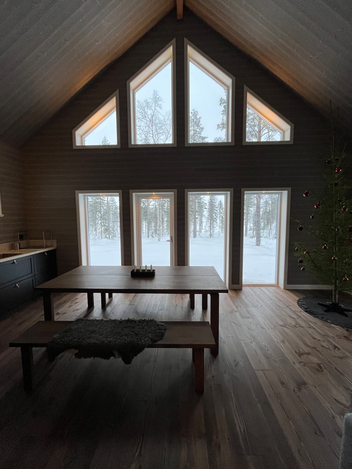 A spacious dining area features a large wooden table surrounded by benches. Expansive windows showcase a snowy landscape outside, allowing natural light to fill the room. A simple kitchen area is visible to the left, while a small decorative tree adds a touch of warmth.