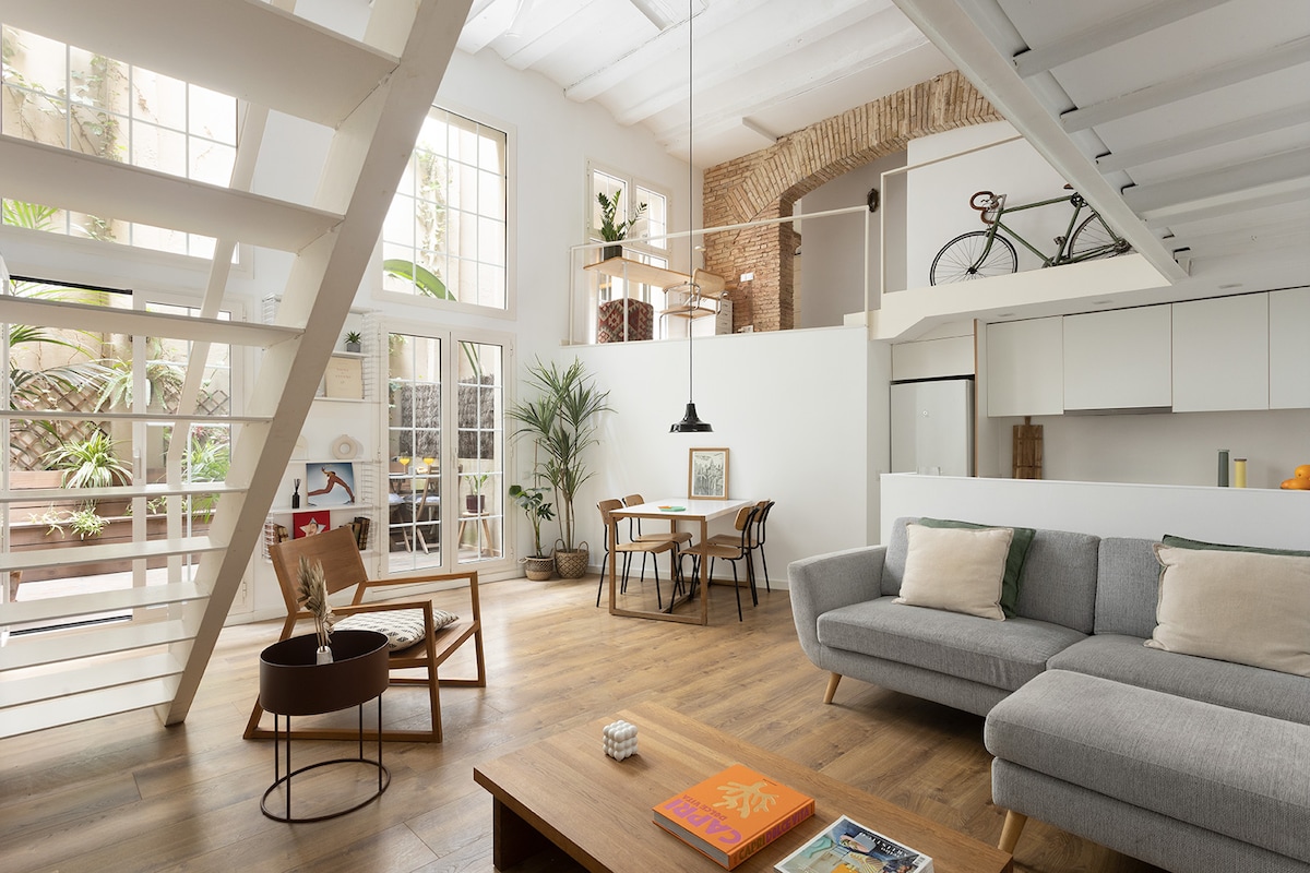 An open-plan living area features high ceilings and large windows, allowing natural light to illuminate the space. A comfortable gray sofa faces a wooden coffee table, while a dining area with a round table and chairs is visible. A potted plant adds a touch of greenery.