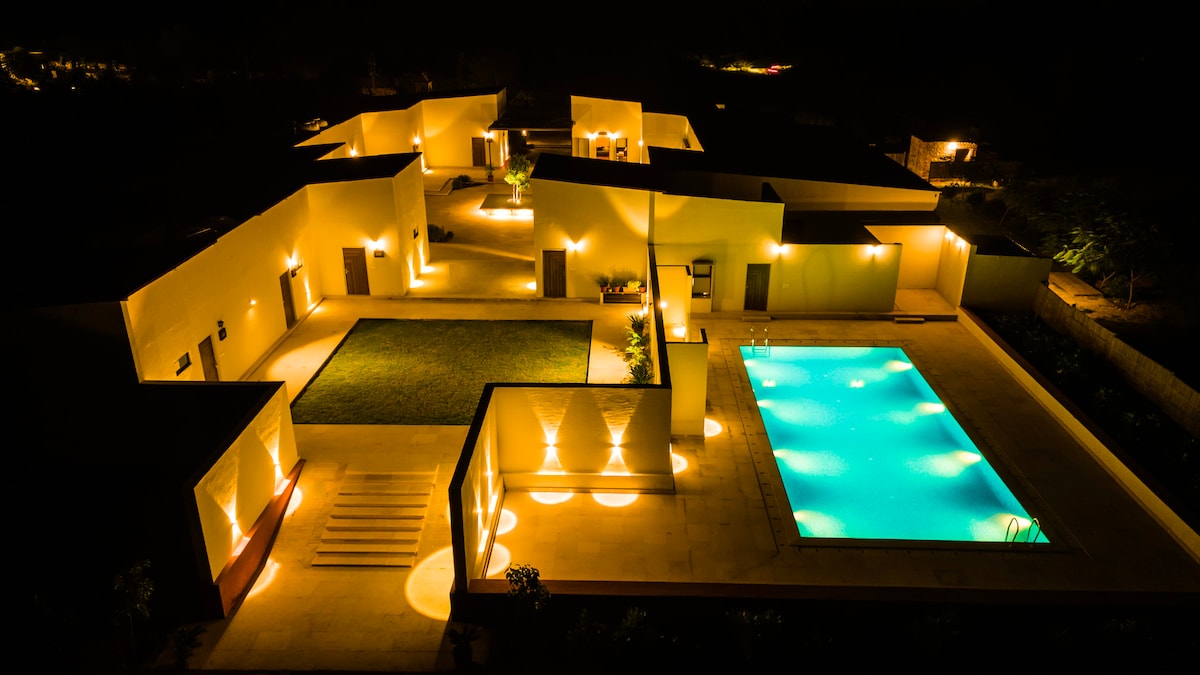 An aerial view of a modern property illuminated at night, showcasing a tranquil swimming pool at the center. Soft lighting enhances the architecture, revealing spacious outdoor areas and manicured landscaping, contributing to a serene atmosphere.