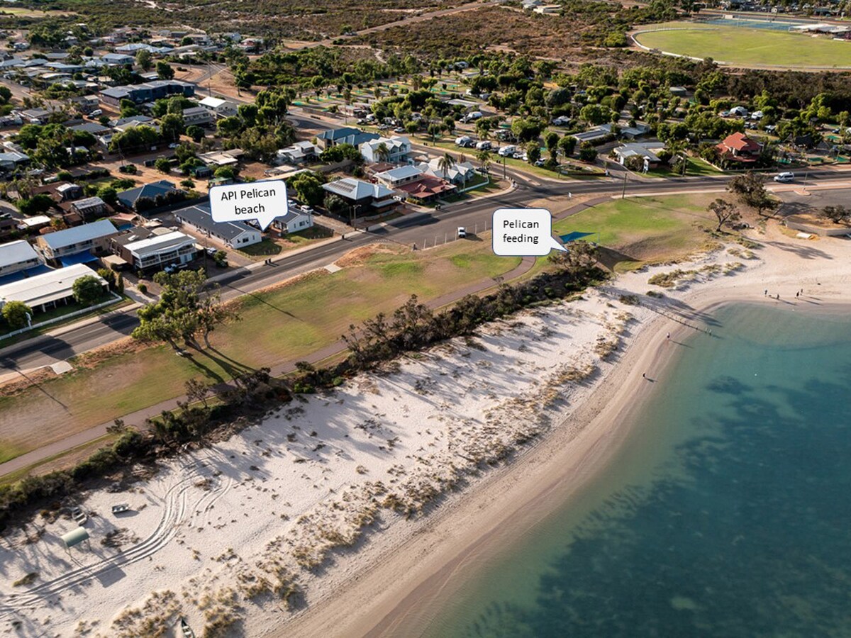 An aerial view captures a coastal area featuring a sandy beach alongside clear waters. The shore is lined with gentle waves, and grassy spaces are visible in the foreground. A small section of the town shows various structures, including residential homes and roads.