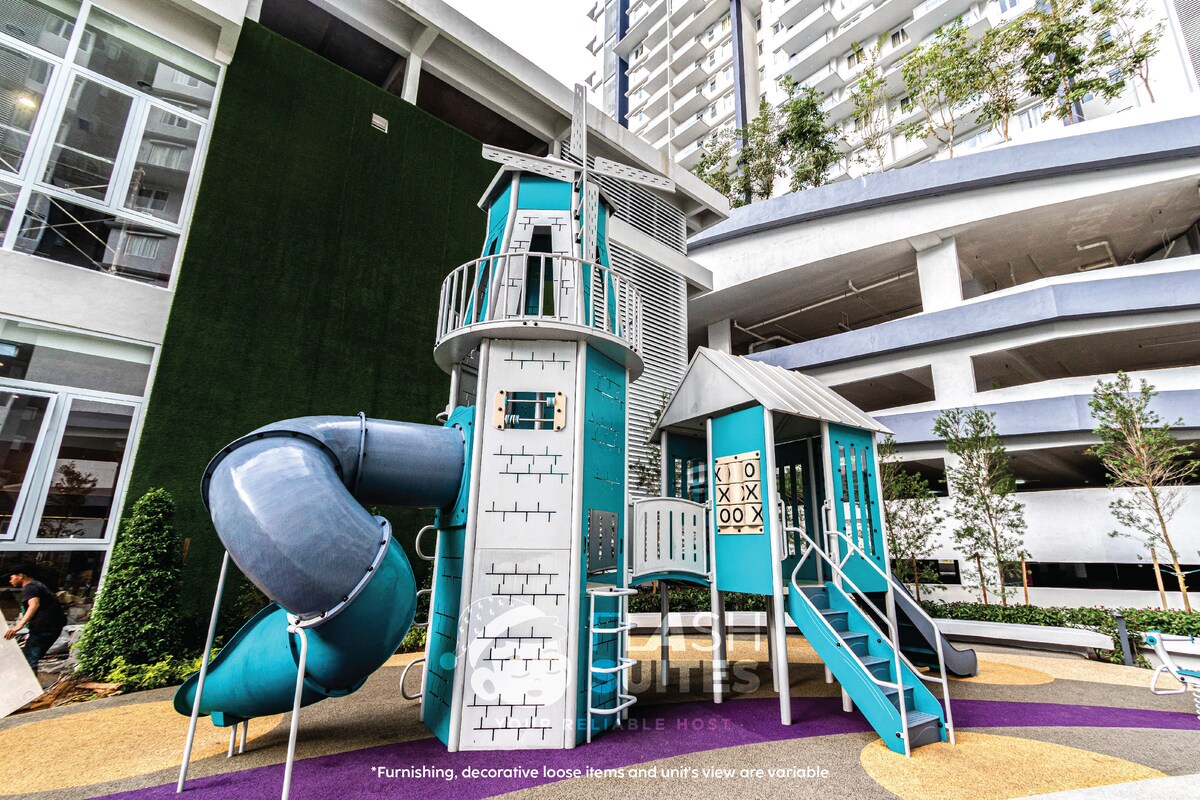 A children's playground is depicted, featuring a multi-level play structure with a blue slide and various climbing elements. The area is landscaped with soft ground covering and surrounded by greenery, offering a safe space for play.